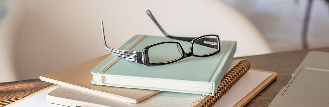 Stack of notebooks and papers on a desk with eyeglasses, a pen, and part of a laptop.